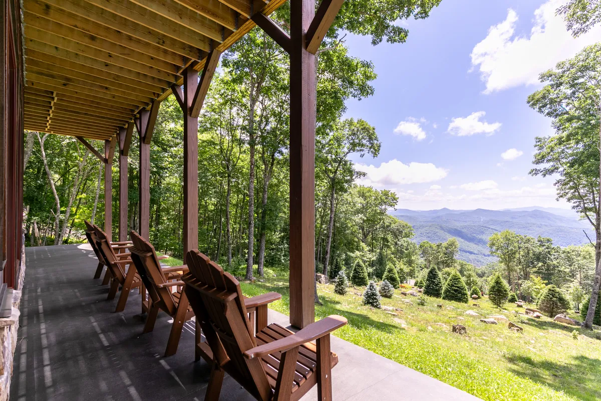 A pair of classic wooden rocking chairs are positioned on the porch, facing an expansive, breathtaking mountain view with lush greenery all around. The scene exudes tranquility and encourages contemplation or socializing while surrounded by nature.