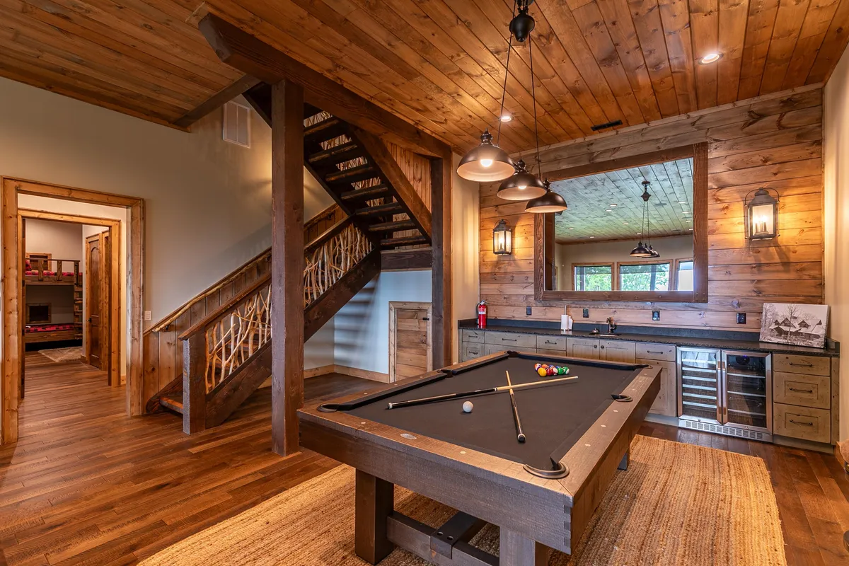 A spacious game room angle highlighting a pool table, a staircase leading to another level, a bar area for drinks, and a large mirror reflecting the room's openness. The design is casual and welcoming, ideal for group entertainment and evenings in.