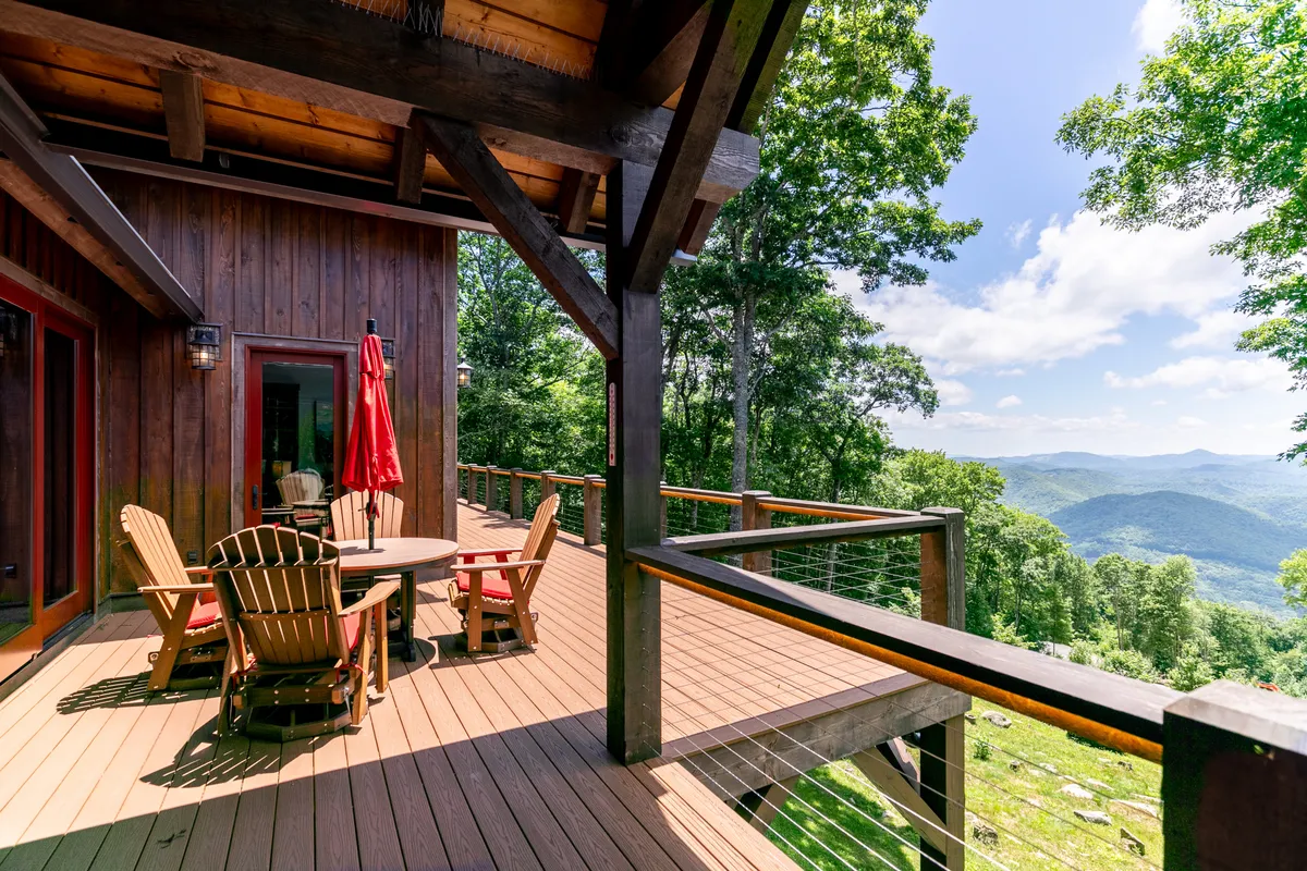 A spacious wooden deck with a table and chairs set beneath a red umbrella, offering an unobstructed view of the surrounding mountains. The setting is designed for relaxation and outdoor dining, with the tranquil atmosphere enhanced by natural greenery and the expansive landscape beyond.