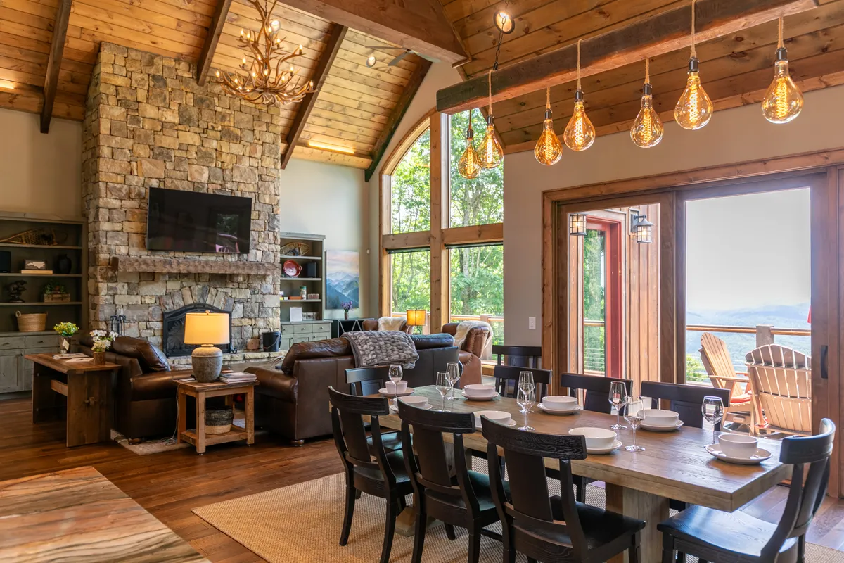 This image beautifully captures the great room’s stacked stone fireplace, comfortable seating area, expansive dining table, and large windows showcasing picturesque mountain views. The chandelier and layout suggest a welcoming space ideal for gatherings and celebrations.