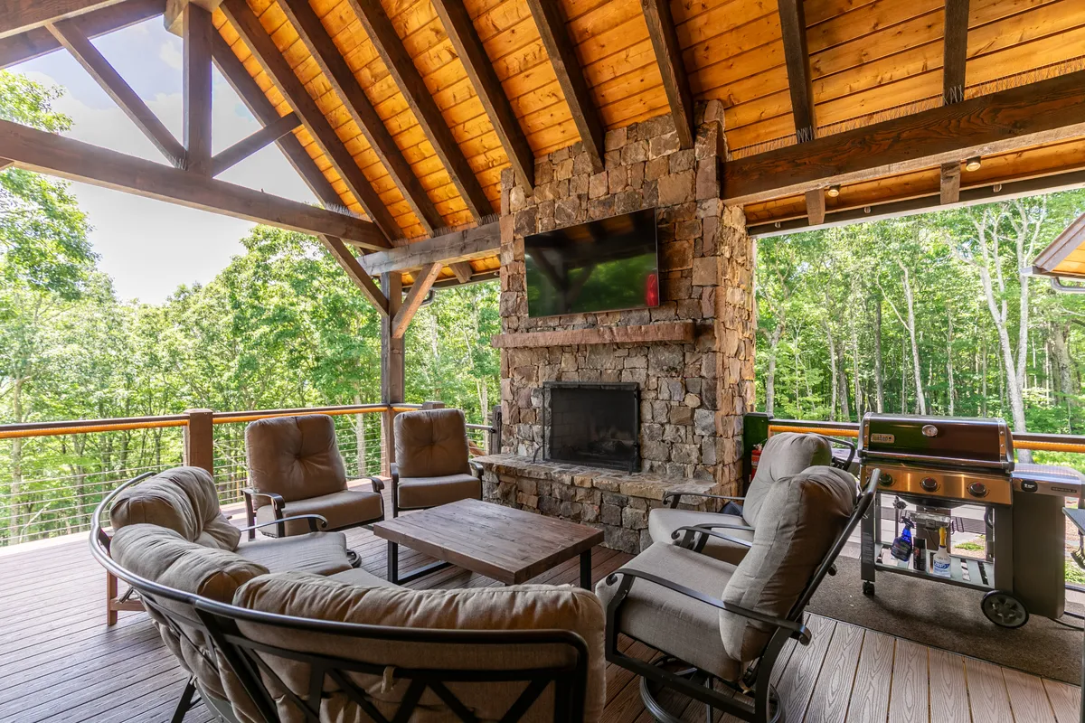 A cozy outdoor living area with a stone fireplace, comfortable seating, and wooden beams, designed for fireside evenings. An outdoor TV is present, perfect for watching the game or gathering with friends and family, creating an inviting alfresco living environment.