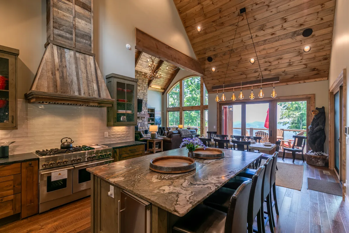 A warm, inviting kitchen scene featuring a large rustic island, modern appliances, and exposed wooden beams. Expansive windows bring the outdoors in, tying the kitchen to the surrounding mountain landscape.
