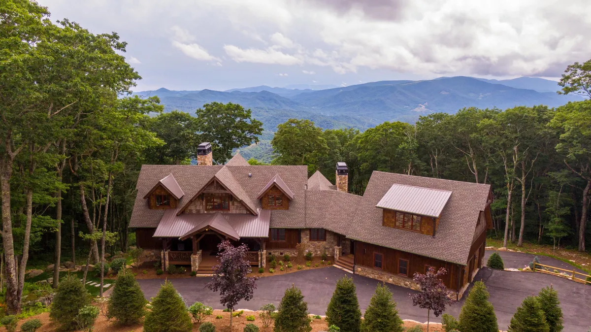 The hero image showcases the grand entrance of a luxury mountain home framed by lush trees, with sweeping views of the Blue Ridge Mountains. The serene scene suggests space for outdoor gatherings, relaxation on the welcoming porch, and panoramic sunset vistas over the ridgeline.