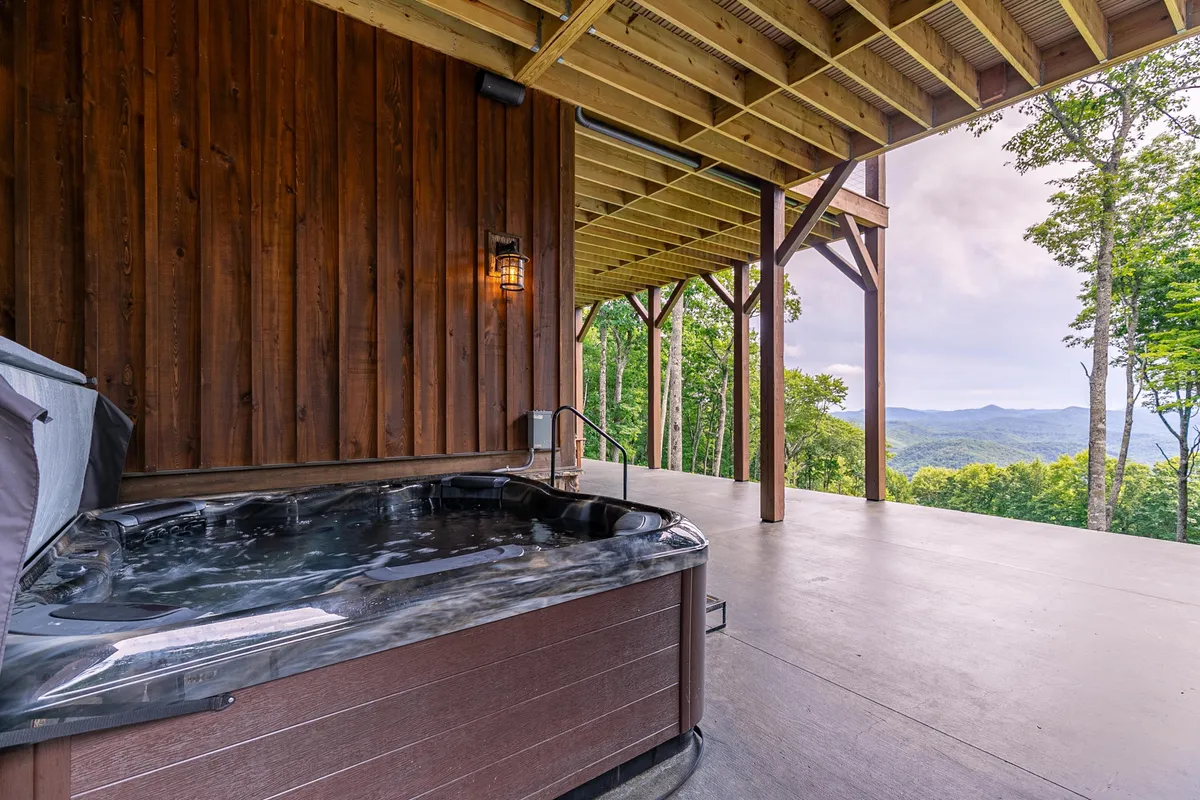 This exterior image features a spacious hot tub set against a wooden wall, with an expansive mountain view in the background. The setting is serene and captures the essence of outdoor relaxation, with natural greenery framing the scene, inviting guests to unwind while taking in the spectacular vistas.