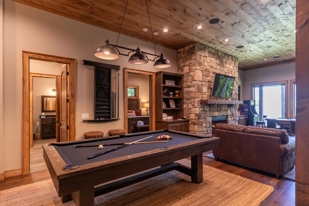 A bright game room featuring a pool table as the centerpiece, backed by a stone fireplace, large windows letting in natural light, a wooden ceiling, and a leather couch for comfortable seating. The friendly setup encourages group activities and relaxation, matching the energetic and social vibe described in the section.