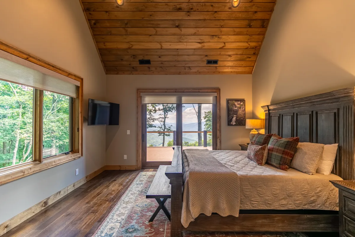 A tranquil king bedroom suite featuring a large bed with decorative pillows, wooden ceiling beams, expansive windows overlooking scenic landscapes, and wooden furniture, all suffused with natural light. The setting exudes calm and showcases plush bedding and inviting mountain views.