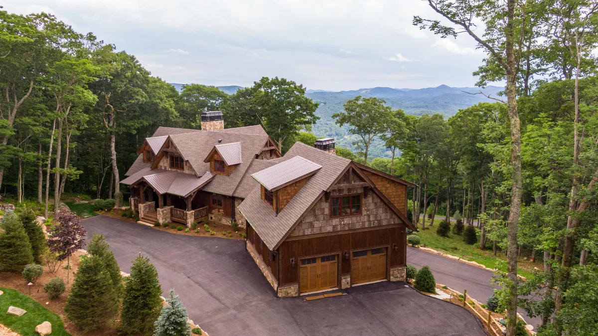 The main entrance of the house, featuring a striking stone façade, surrounded by mature trees and set against a backdrop of rolling mountains. The inviting porch and natural setting capture the connection between the home and its stunning environment.