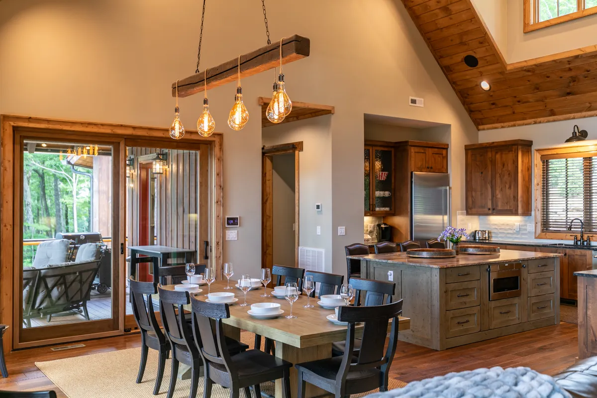 This photo centers on the dining area and kitchen island, with large windows allowing natural light to fill the space. The image highlights the roomy dining table and the gourmet kitchen setup, including hanging light fixtures and plenty of seating for communal meals.